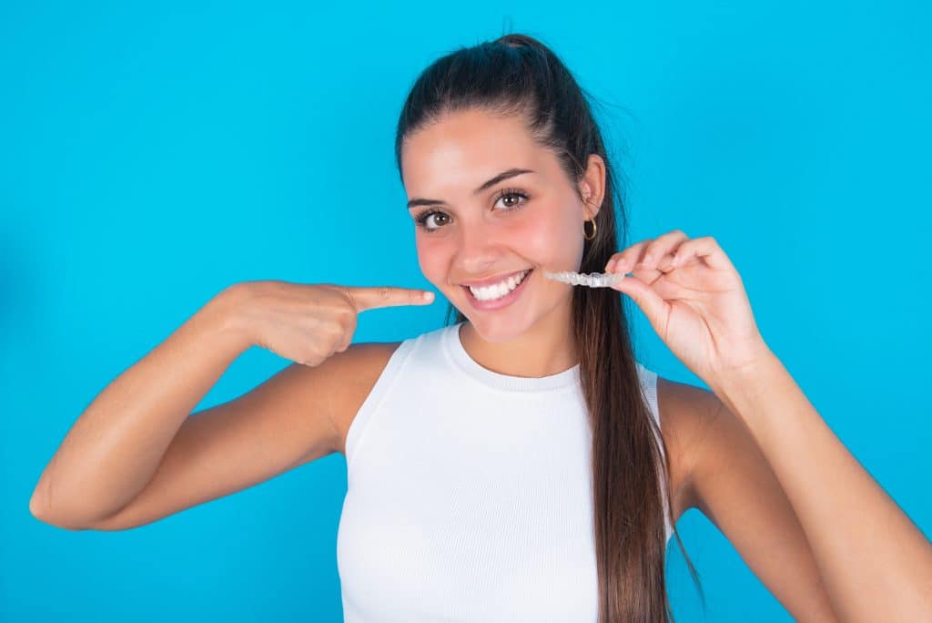 beautiful brunette woman wearing white tank top over blue background holding an invisible aligner and pointing to her perfect straight teeth. Dental healthcare and confidence concept.