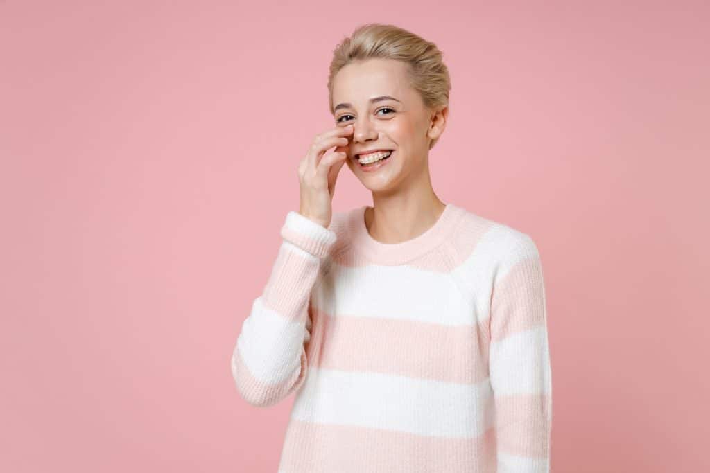 Young smiling blonde caucasian woman 20s short haircut dental braces nude makeup in casual knitted sweater touching face soft skin nose look camera isolated on pastel pink background studio portrait.